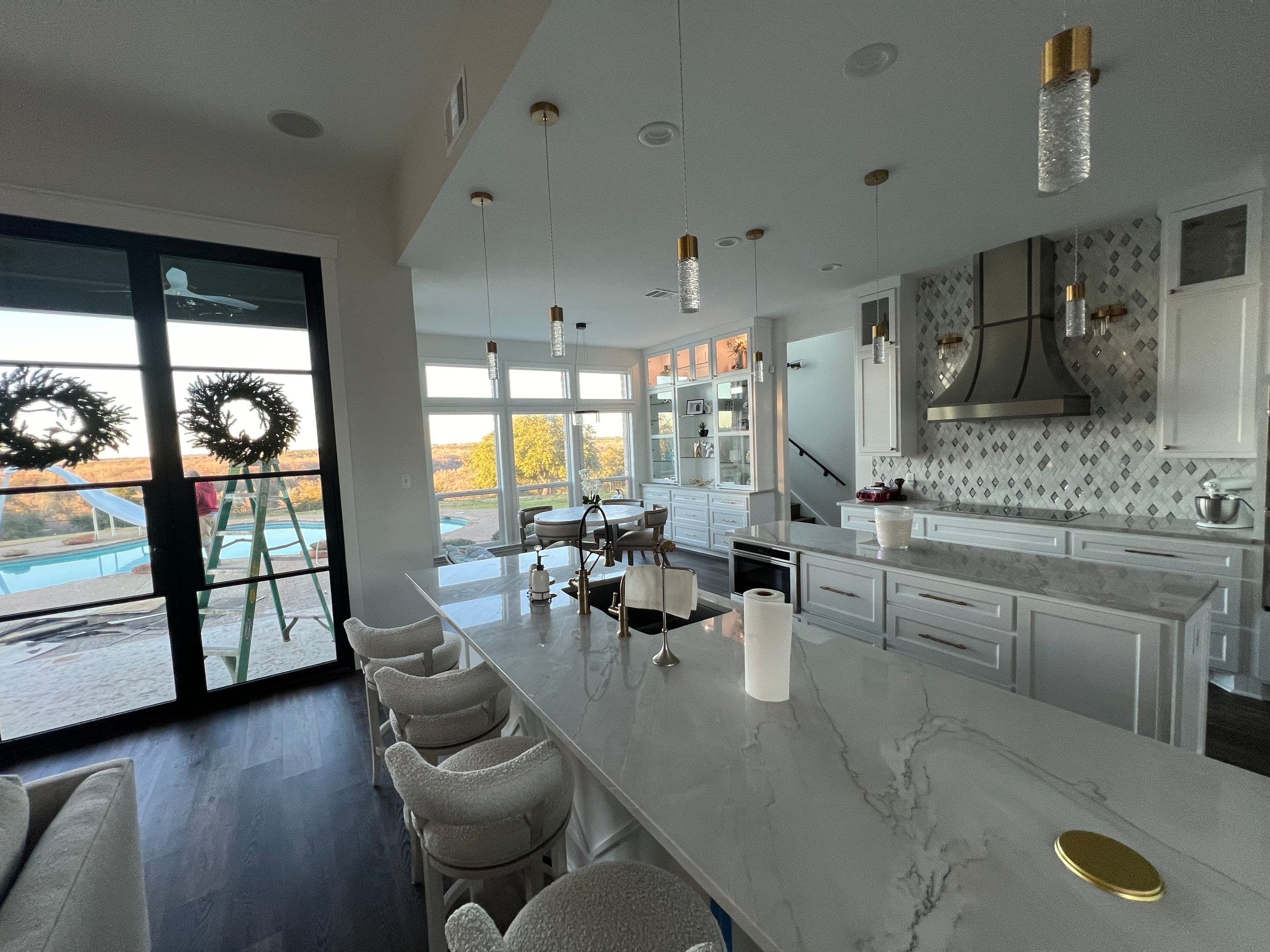 Modern white kitchen with marble island, gold pendant lights, and glass doors overlooking a pool.